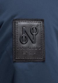 Zwarte leren label met een reliëf "N" logo en de tekst "NAVAHOO EST 2014" op een donkerblauwe stofachtergrond, met een getextureerd oppervlak en stiksel.