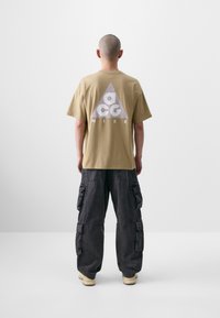 Beige t-shirt i bomuld med et stort grått trekantet logo med teksten "ACG" og "NIKE". Kombineret med mørkegrå cargo bukser og beige sko.