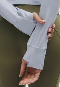 Osoba wearing světle modré rukávy s otvory pro palce, ruce zkřížené před olivovými legínami.