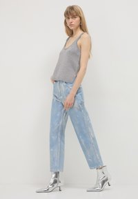 Giovane donna in piedi che indossa un top in maglia grigia, jeans azzurri con effetto acid washed e stivali neri alla caviglia su uno sfondo chiaro.
