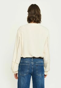 Ljuskrämfärgad blus med samlade långa ärmar, har delikat sydda detaljer längs axelsömmarna, kombinerad med blå denimjeans.