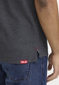 Osoba v tmavě šedém tričku s červenou nášivkou ISOLID na lemu, v modrých džínách, s uvolněnou paží podél těla.