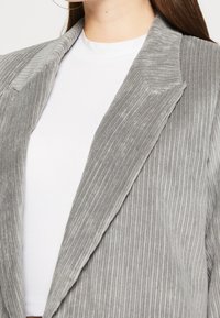 Blazer in velluto grigio con righe verticali pronunciate, revers a punta e texture morbida, indossato sopra una maglia bianca.