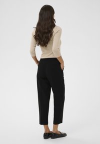 Haut beige ajusté associé à un pantalon noir décontracté. Le pantalon présente des poches latérales et une texture lisse, se terminant au-dessus de la cheville avec des chaussures noires élégantes.