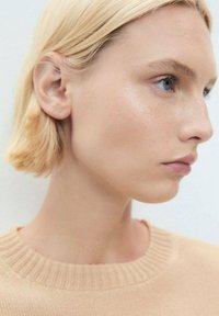 Portrait of a person with short, straight blonde hair, wearing a light beige sweater with a ribbed neckline, smooth skin, and neutral expression.