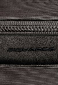 Reistas van donkergrijze stof met een geaccentueerd leeraccent, een metalen ritssluiting en subtiele, in reliëf aangebrachte merknaam dicht bij de ritssluiting.