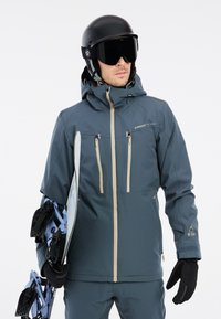 Skijacke in Dunkelblau mit beigen Reißverschlüssen, Kapuze und Belüftungslöchern; kombiniert mit einem schwarzen Helm, Skibrille und Snowboardbindungen.