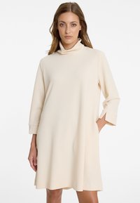 Frau mit hellbraunem Haar, die ein knielanges, cremefarbenes Kleid mit hohem Kragen und Dreiviertelärmeln trägt, eine Hand in der Tasche.