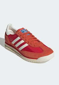 Zapatilla roja y naranja con parte superior de ante y nailon, tres rayas blancas y un collar acolchado. Presenta una suela de goma texturizada.