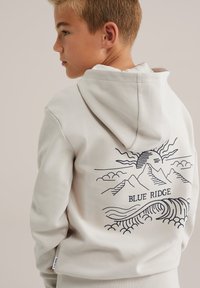 Crème hoodie met een grafisch ontwerp van bergen en golven op de achterkant, met de tekst "BLUE RIDGE", gemaakt van zachte stof.