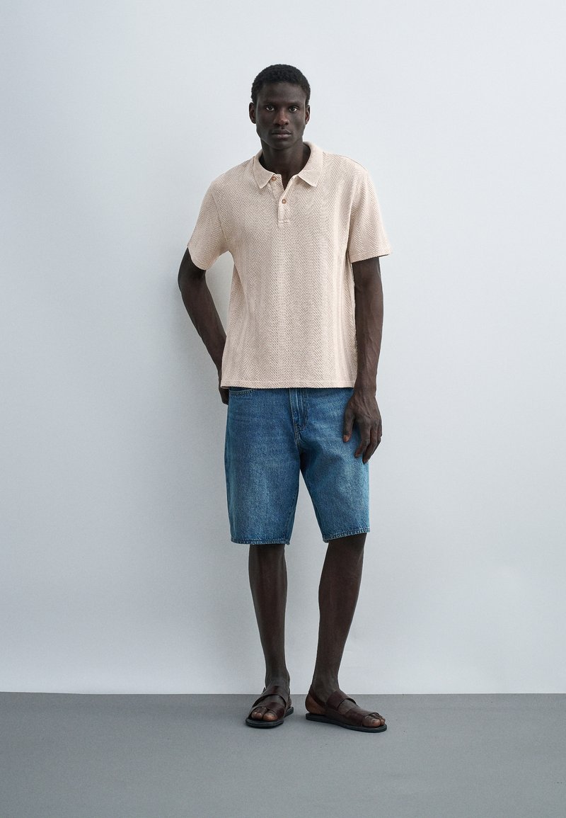 Homme portant un polo beige à manches courtes, un short en jean bleu et des sandales marron, debout devant un mur gris clair uni.