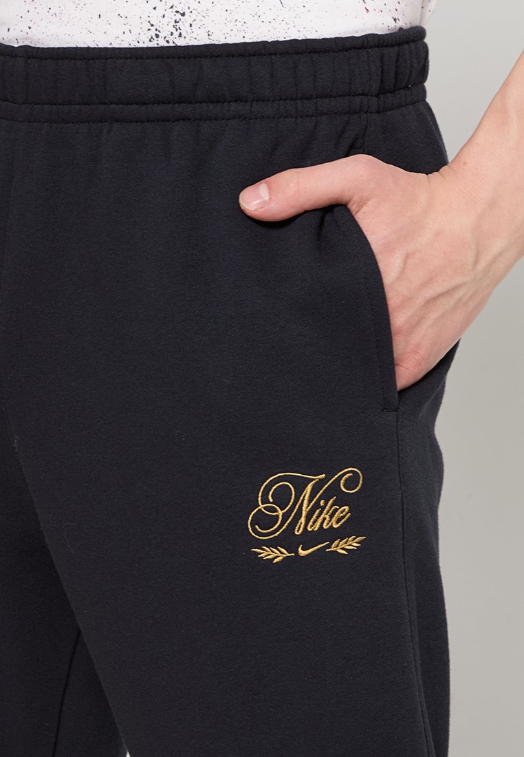 Schwarze Sweatpants mit einer weichen Textur, die ein goldgesticktes Nike-Logo am linken Oberschenkel und einen elastischen Bund aufweisen.