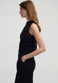 Top senza maniche lavorato a maglia nera con design testurizzato e motivo aperto. Presenta un collo alto ed è abbinato a una lunga gonna nera.