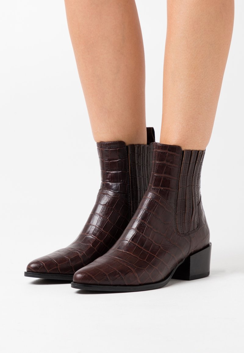 Bottines marron foncé à texture crocodile, bouts pointus et talons blocs bas portées sur jambes nues sur fond blanc.