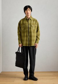 Chemise écossaise verte à deux poches sur la poitrine, associée à un pantalon noir et un sac fourre-tout noir. La chemise présente un design à boutons et des manches longues.