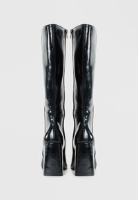 Botas negras hasta la rodilla con un acabado de cuero brillante, un tacón cuadrado y cremalleras laterales para facilitar su uso. Textura suave con un diseño minimalista.