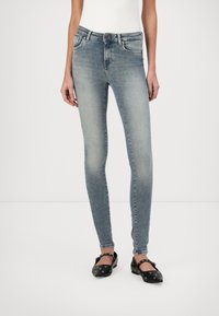 Vaqueros ajustados azul descolorido con cintura de tiro medio, cinco bolsillos y trabilla para cinturón. Combinados con zapatos planos negros con detalles circulares.