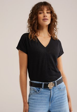 Žena u crnoj V-izrez majici i svijetloplavim trapericama s crnim remenom s zlatnom kopčom u obliku školjke, stoji na jednobojnoj pozadini.