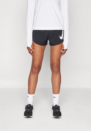 Person, die schwarze Nike-Laufshorts, ein weißes langärmeliges Oberteil, weiße Nike-Socken und schwarze Sportschuhe trägt und vor einem einfarbigen Hintergrund steht.