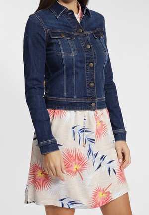 Vrouw met een donkerblauw spijkerjasje over een lichte jurk met een roze, geel en blauw bloemenpatroon, staand tegen een effen achtergrond.