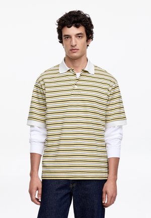 Jaunas vyras dėvi geltoną, baltą ir juodą horizontalios juostelės polo marškinėlį ant baltos ilgarankovės marškinėlių ir tamsiai mėlynus džinsus, stovintis ramiai.