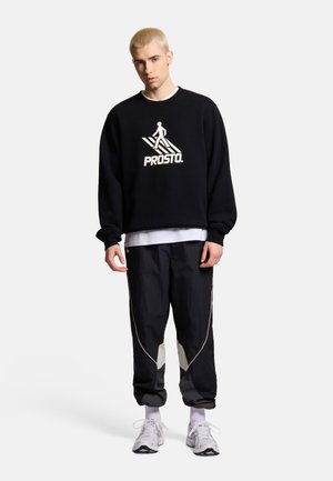Młody mężczyzna o krótkich blond włosach, ubrany w czarną bluzę Prosto, czarne spodnie dresowe z białymi łatami, białe trampki i białe skarpetki.