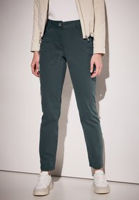 Pantalones de algodón verde oscuro, ajustados, con una textura suave, que cuentan con un cierre frontal con botón y un clásico diseño de cinco bolsillos.
