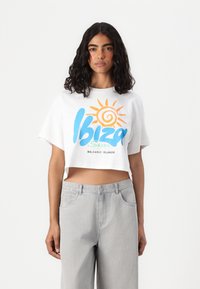 Κοντομάνικο λευκό μπλουζάκι με πολύχρωμο γραφικό "Ίμπιζα Ήλιος" και εικονίδιο ήλιου. Συνδυάζεται με ανοιχτό γκρι ψηλόμεσο τζιν. Καθημερινός σχεδιασμός.