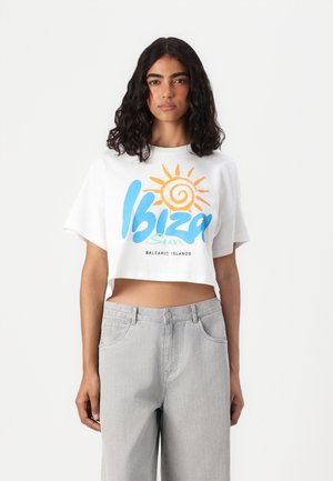 Valkoinen t-paita, jossa on värikäs "Ibiza Sun" -grafiikka auringon ikonilla. Yhdistetty vaaleanharmaisiin korkeavyötäröisiin farkkuihin. Rentoa muotoilua.