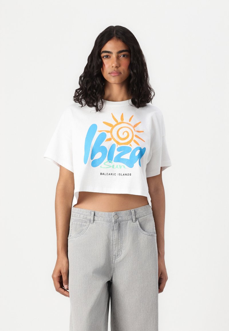 Κοντομάνικο λευκό μπλουζάκι με πολύχρωμο γραφικό "Ίμπιζα Ήλιος" και εικονίδιο ήλιου. Συνδυάζεται με ανοιχτό γκρι ψηλόμεσο τζιν. Καθημερινός σχεδιασμός.