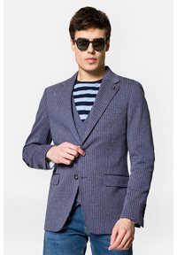 Granatowa i biała marynarka w kratę z szpiczastymi klapami, na pojedynczy guzik i dwoma przednimi kieszeniami, w połączeniu z pasiaste koszulą pod spodem.