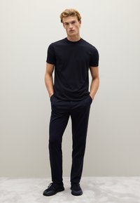 Camiseta de algodón azul marino con cuello redondo, combinada con pantalones oscuros. El modelo está de pie con las manos en los bolsillos sobre una alfombra gris claro.