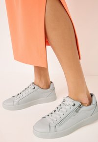 Baskets en cuir gris avec bouts ronds, semelles plates et lacets. Comprennent une fermeture éclair sur le côté et des détails de couture. Portées avec une jupe de couleur corail.