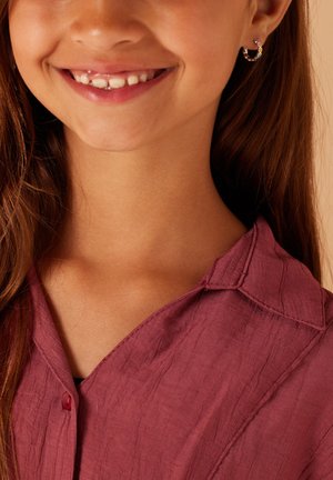 Jongere in een button-down overhemd met een gekreukt textuur in een diepe bordeauxkleur, vergezeld van kleine ringoorbellen met gekleurde accenten.