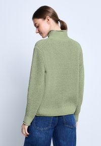 Groene gebreide trui met een hoge kraag, gestructureerd materiaal en lange mouwen. Draagt met blauwe jeans, die een ontspannen pasvorm en een geribbelde onderrand tonen.