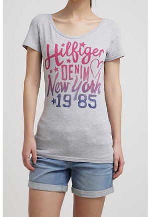 Kobieta w szarej koszulce z napisem "Hilfiger Denim New York 1985" i niebieskich, podwiniętych szortach dżinsowych, stojąca na jednolitym tle.