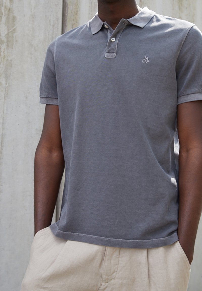 Szara koszulka polo z miękkiej tkaniny, z dwuknapowym dekoltem, krótkimi rękawami i małym logo na piersi. W zestawie z jasnymi spodniami.