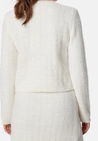 Vit, strukturerad jacka med kort design och långa ärmar, som har ett fyrkantigt mönster och rund halsringning, kombinerad med en matchande kjol.