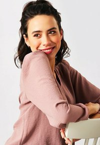 Blusa rosa chiaro, con texture e maniche lunghe, indossata da una persona con il braccio appoggiato su una sedia. Tessuto morbido con una vestibilità comoda.
