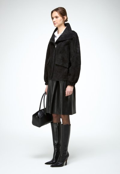 Femme portant une veste noire zippée, une jupe en cuir noir, des bottes noires montant jusqu'aux genoux, tenant un sac à main noir, debout de profil sur un fond blanc.