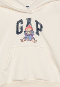 Hanorac crem cu buzunar frontal, litere mari "GAP" și o ilustrație cu un urs îmbrăcat într-o haină albastră și o pălărie roșie.