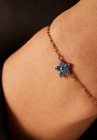 Gouden kettingarmband met een bloemvormig ontwerp, versierd met zes blauwe edelstenen en een centrale gouden kraal. Gladde textuur op de huid.