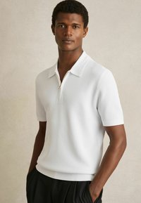 Mann mit kurzen, lockigen Haaren, der ein weißes Kurzarm-Strickpolo mit Reißverschluss trägt und mit den Händen in den Taschen seiner schwarzen Hose vor einem schlichten Hintergrund steht.