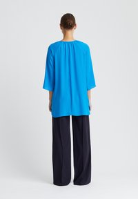 Helderblauw, lichtgewicht blouse met driekwart mouwen en geplooid achterkantdetail, gecombineerd met donkere, brede broekspijpen. Eenvoudig ontwerp.