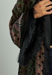 Kimono multicolor cu un model floral verde și maro, împodobit cu franjuri negri intricate pe mâneci și cu o textură netedă.