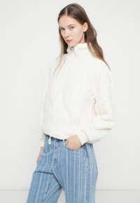 Biely flísový pulóver s vysokým golierom, textúrovaným povrchom a rebrovanými manžetami. Kombinovaný so svetlomodrými pruhovanými džínsami.