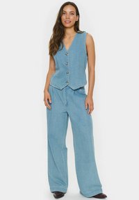 Svetlo modra denimka z petimi gumbi in V-izrezom, v kombinaciji z širokimi hlačami iz istega materiala. Gladka tekstura, sproščen kroj.
