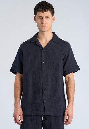 Jonge man draagt een donkerblauw gestructureerd overhemd met korte mouwen en knopen, staand voor een effen lichte achtergrond.