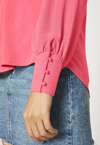 Hand, die ein pinkfarbenes Langarmshirt mit stoffüberzogenen Knöpfen am Bündchen trägt, kombiniert mit einem blauen Jeansrock.