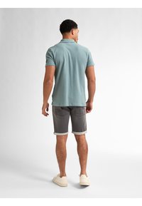 Camisa polo azul claro de manga corta, combinada con pantalones cortos grises con dobladillo. Modelo con zapatillas crema, visto desde atrás.